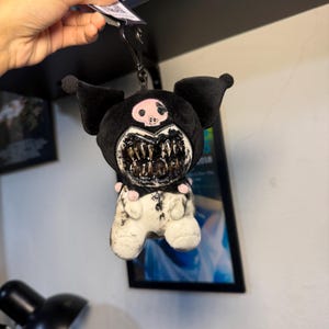 Puede incluir: Un llavero de peluche con un personaje negro de orejas puntiagudas, un emblema de calavera rosa en la frente y una boca grande y abierta llena de dientes afilados y realistas. El cuerpo es blanco con marcas negras desgastadas y detalles rosas.
