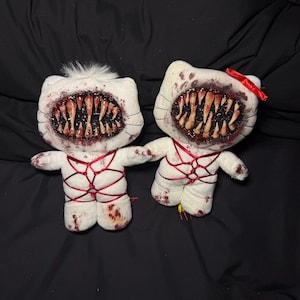 May include: Two white plush cat-like dolls featuring large, gaping mouths filled with sharp, realistic teeth. The dolls are splattered with red stains and wrapped in thin red ribbon. One doll has a small tuft of white fur, the other a red bow.