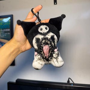 May include: A plush keyring featuring a black and white character with a jester-style hood, a skull emblem on the forehead, and a wide, gaping mouth filled with sharp, jagged teeth and a long, protruding pink tongue, finished with a black clip.