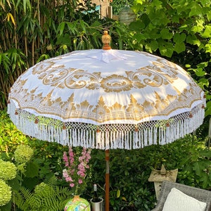 May include: A white and gold patterned patio umbrella with white fringe, a wicker chair with a white cushion, a small table with a decorative vase and a book, and green plants in the background.