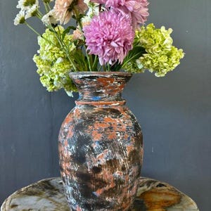 Può includere: Un vaso in ceramica rustico, dipinto a mano con una finitura usurata in tonalità di nero, arancione e blu. Il vaso è pieno di un bouquet di fiori rosa, bianchi e verdi.