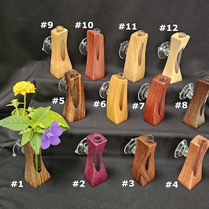 May include: Twelve wooden wall-mounted bud vases with glass inserts and suction cups, numbered 1 through 12. Each vase features a unique wood grain and color, ranging from light tan to dark brown and purple. One vase holds a yellow and purple flower.