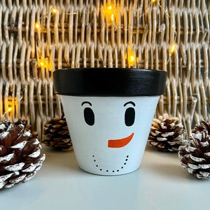 Pode incluir: Pote de cerâmica branco com borda preta, pintado para parecer um boneco de neve com olhos pretos, um sorriso laranja e pontos brancos para botões. O pote está sobre uma superfície branca com pinhas.