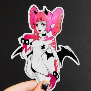 Peut inclure: Autocollant repr&eacute;sentant une illustration de d&eacute;mon f&eacute;minin avec des cheveux et des accents roses. Le d&eacute;mon a des ailes noires, un collier et des menottes. Le design incorpore un m&eacute;lange de couleurs noir, blanc et rose.