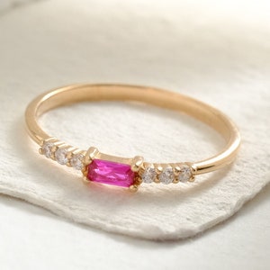 May include: A gold ring featuring a central rectangular baguette-cut pink gemstone flanked by three small round clear stones on each side. The band is thin and polished, showcasing a delicate design with a vibrant pink center stone and clear accents.