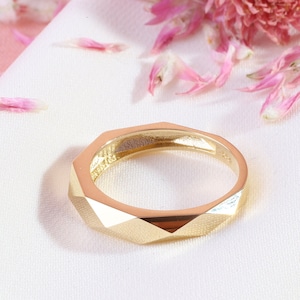 May include: A gold geometric ring with a faceted design. The ring is on a white and pink fabric background.