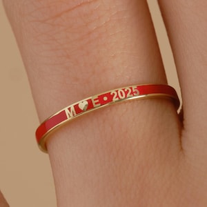 May include: A gold ring with a red enamel band. The ring features the inscription "M♡E 2025". The ring is a close-up shot, highlighting the details of the inscription and the band's color.