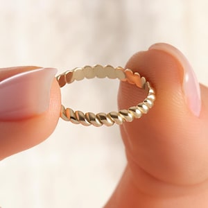 May include: A gold-colored ring with a twisted rope design. The ring is held between two fingers, showcasing its delicate details. The ring's texture is visible, highlighting the craftsmanship. The ring is a piece of jewelry.