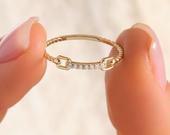 May include: A delicate gold ring with a unique design. The ring features a series of small, sparkling stones set in a straight line, with decorative elements on either side. The ring is held between two fingers.