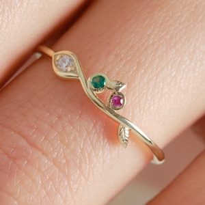 Peut inclure: Une bague en or délicate avec un motif de feuille et de vigne. La bague est ornée de trois pierres précieuses : une claire, une verte et une rouge. La bande de la bague est fine et élégante, parfaite pour un usage quotidien.