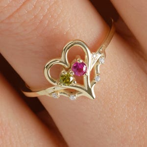 May include: A gold ring with a heart-shaped design. It features a pink gemstone and a green gemstone, with small white accents. The ring is worn on a finger.