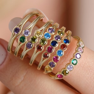 May include: A collection of gold-colored rings, each adorned with a row of colorful gemstones. The gemstones include purple, green, yellow, blue, and pink. The rings are displayed on a finger, showcasing their design and color variations.