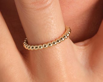 May include: A gold beaded ring is worn on a finger. The ring is made of small, round beads that are closely set together, creating a textured surface. The ring's color is a bright, shiny gold.