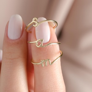 May include: Three gold rings, each with a cursive letter: "S", "a", and "m". The rings are displayed on a finger, highlighting their minimalist design and elegant script. The rings are a warm gold colour.