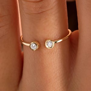 May include: A delicate gold ring featuring two small, round, clear gemstones set in open bezels. The ring's band is thin and polished, with a simple, elegant design. The gemstones are evenly spaced, creating a minimalist and modern aesthetic.