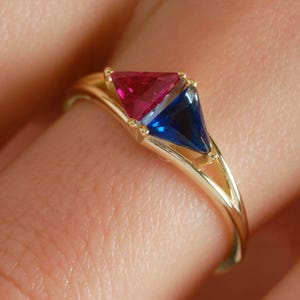 May include: A gold ring featuring two triangular gemstones, one ruby red and the other sapphire blue. The ring's band has a delicate, open design. The gemstones are set close together, creating a striking contrast of colors.