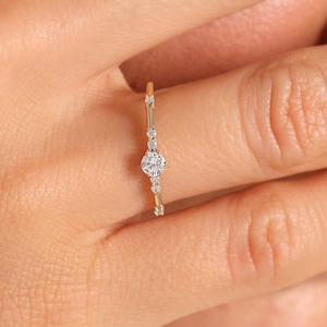 May include: A delicate gold ring with a central round diamond and smaller diamonds and gold accents. The ring is set on a thin band and is shown on a finger.