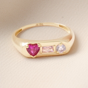 May include: A gold ring with a rectangular face, featuring a heart-shaped pink gemstone, a rectangular pink gemstone, and a round, light purple gemstone. The ring is set against a neutral background.