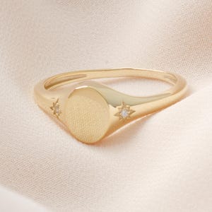 May include: A gold-colored signet ring with an oval face and starburst accents. The ring has a smooth band and a textured oval center. Small, clear gemstones are set in the starburst designs.