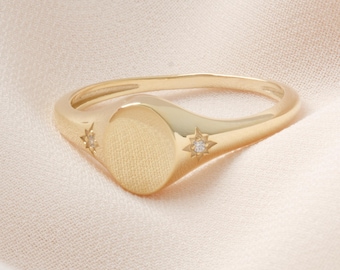 May include: A gold-colored signet ring with an oval face and starburst accents. The ring has a smooth band and a textured oval center. Small, clear gemstones are set in the starburst designs.