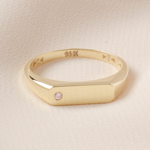 May include: A gold ring with a rectangular face and a small pink gemstone. The ring is marked with "14K" and has a polished finish. The ring is set on a cream-colored fabric.