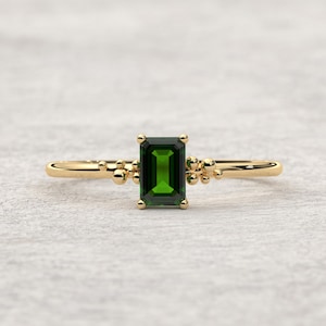 May include: A gold ring featuring a green emerald-cut gemstone and small gold beads on the band.