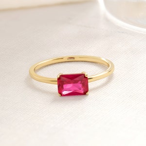 May include: A gold ring with a rectangular, bright pink gemstone. The ring band is thin and smooth, with the inscription "MIN14K" visible. The gemstone is set in a simple, elegant design, showcasing its vibrant color.