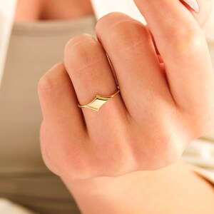 May include: A gold ring with a geometric, diamond-shaped design. The ring is worn on a finger.
