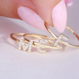 Peut inclure: Trois bagues dorées avec les lettres "M", "C" et "E" incrustées de petits diamants transparents. Les bagues sont présentées sur une surface en satin blanc, avec un fond rose. Les bagues sont tenues par une personne.