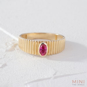 May include: A gold ring with a pink oval gemstone set in the centre. The band of the ring has a textured, ribbed design.