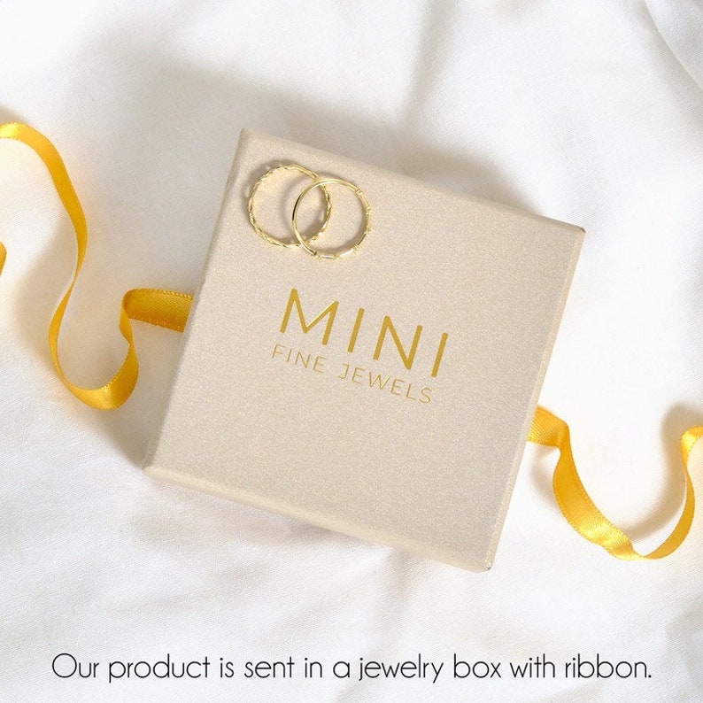 Op de afbeelding: Een beige juwelendoos met een gouden lint en twee gouden ringen. Op de doos staat "MINI FINE JEWELS" gedrukt.