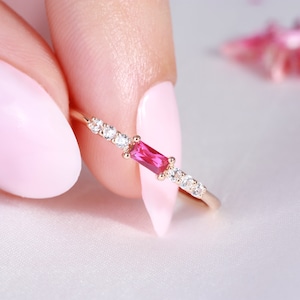 May include: A delicate gold ring featuring a rectangular, vibrant pink gemstone at the center, flanked by smaller, clear gemstones. The ring is set against a soft, white background, highlighting its elegant design.