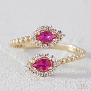 May include: A gold ring with two pear-shaped pink gemstones surrounded by smaller clear gemstones. The ring is beaded and has a delicate design.