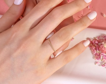 May include: A delicate gold chain ring on a hand with white nail polish. The hand is resting on a white surface with pink flower petals scattered around it.
