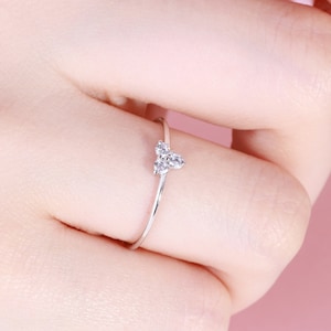 May include: A delicate silver ring featuring a cluster of three small, clear gemstones in a heart-like arrangement. The ring is shown on a finger against a soft pink background, highlighting its minimalist design and sparkle.