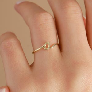 May include: A thin gold ring featuring a lowercase letter 'a' in a cursive script font. The band is smooth and polished, with the letter integrated into the center of the ring, creating a delicate and continuous design across the finger.