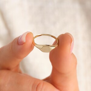 May include: A gold signet ring with a rectangular face is held between two fingers. The ring has a smooth, polished finish and a simple, elegant design. The ring is a classic piece of jewelry.
