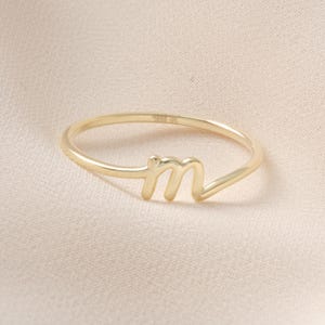 May include: A delicate gold ring featuring the lowercase letter "m" in a cursive style. The ring has a thin band and the letter is slightly raised. The ring is set against a soft, neutral background.
