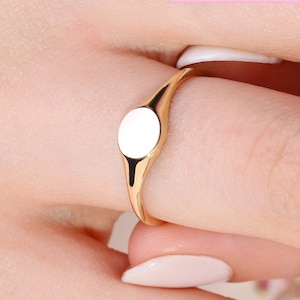 Peut inclure: Une bague signet en or avec un dessus ovale blanc. La bague est portée à un doigt.