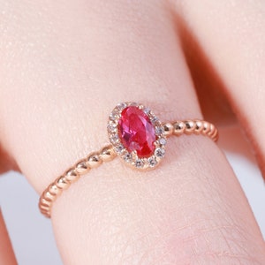 May include: A rose gold ring featuring a central oval-cut vibrant red gemstone surrounded by a halo of small clear stones. The band of the ring has a distinct beaded or textured design, providing a unique look to the metal setting.