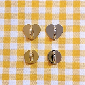 May include: Four gold-colored heart-shaped metal clasps with a ring in the center. The clasps are arranged in a square pattern on a yellow and white checkered background.