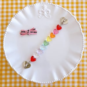 May include: Rainbow heart charm necklace with silver heart clasps. The necklace is made of clear plastic hearts in shades of red, orange, yellow, green, blue, indigo, and violet.