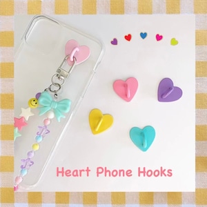 May include: A set of six heart-shaped phone hooks in various colors, including pink, purple, yellow, and blue. The hooks are designed to attach to a phone case and can be used to hang keys, earphones, or other small items. The image also shows a phone case with a beaded charm attached.