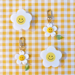 May include: Three white daisy-shaped keychains with smiley faces. Each keychain has a gold metal ring and a small green leaf charm. The keychains are on a yellow and white checkered background.