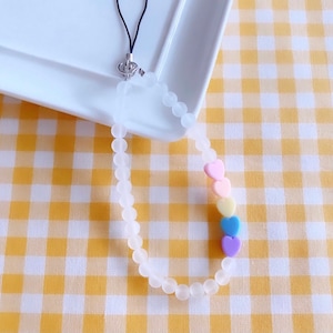 May include: A white beaded phone charm with a rainbow of heart-shaped beads. The charm is attached to a black phone strap.