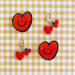 May include: Two red heart-shaped keychains with smiley faces. One keychain has a gold chain and red berry charms. The other keychain has a silver chain and red berry charms.