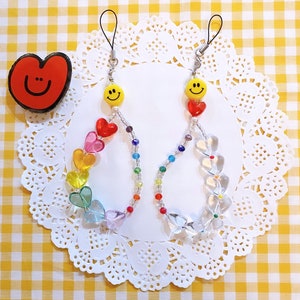 May include: Two rainbow heart charm phone straps with smiley face charms. The charms are attached to a silver chain with a clear heart charm at the bottom.