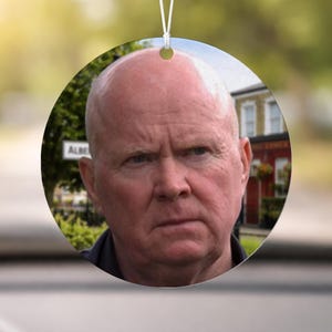 May include: A round, white ceramic ornament featuring a portrait of a man with a bald head and a stern expression. The background includes a street sign that reads "ALBE" and a building with a red facade. A white string is attached for hanging.