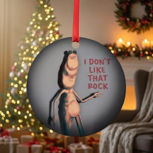 May include: A round Christmas ornament with a red ribbon. The ornament features a cartoon character with the text "I DON'T LIKE THAT ROCK". The character is light brown with a long neck and is pointing to the right.