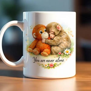 Puede incluir: Taza de cerámica blanca con una ilustración de dos monos abrazándose, uno naranja y otro marrón, rodeados de flores. El texto "You are never alone" está escrito debajo.
